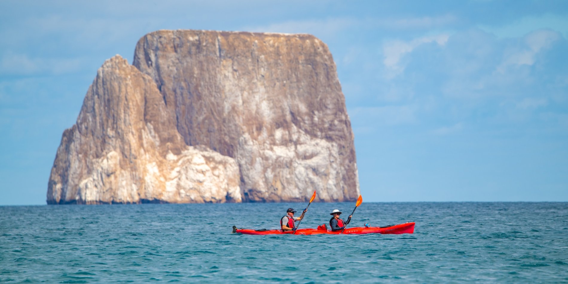 A sea kayak floating in the ocean with a large rock formation behind.