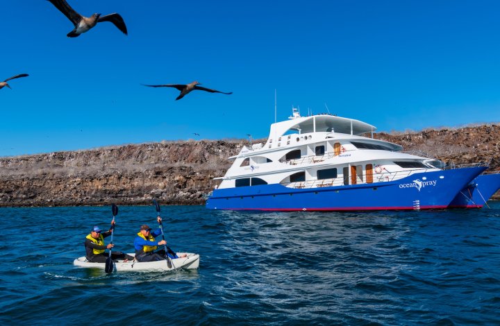Tandem sea kayak with people paddling past the Ocean Spray small cruise ship in the Galapagos