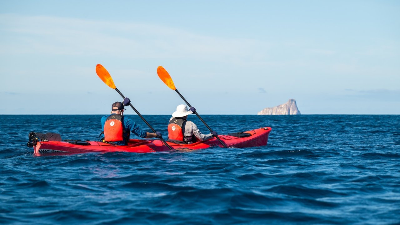 Two kayakers paddling in open ocean waters off the Galapagos Islands with Kicker Rock visible in the distance.