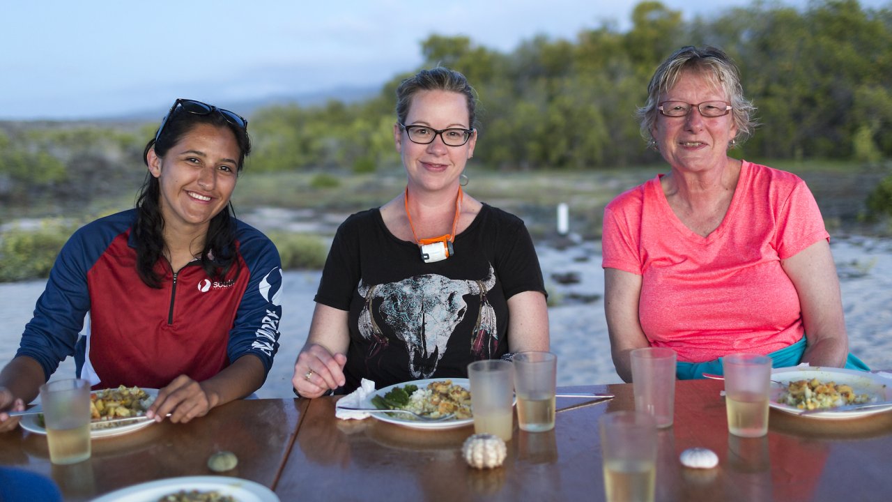 Three ladies on a beach eating dinner at a table while camping in the Galapagos