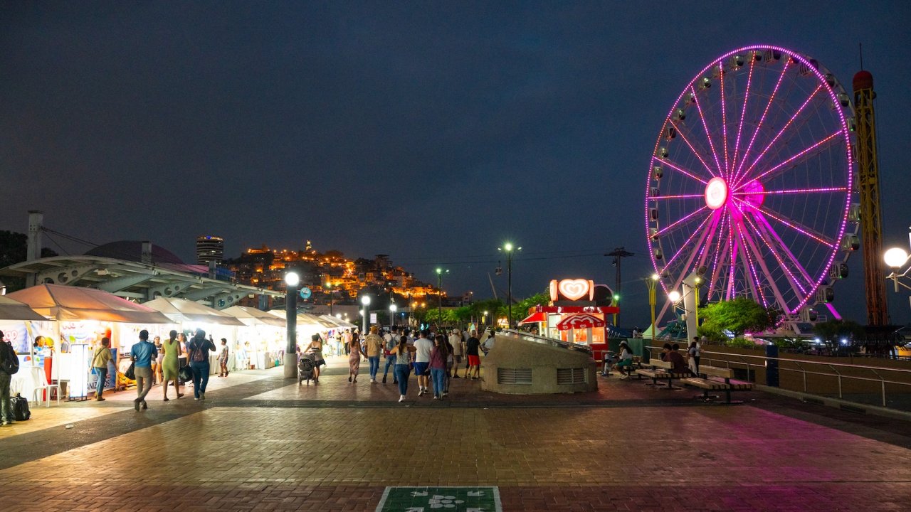 Families exploring a lively waterfront market at night with a glowing Ferris wheel during a Galapagos trip.
