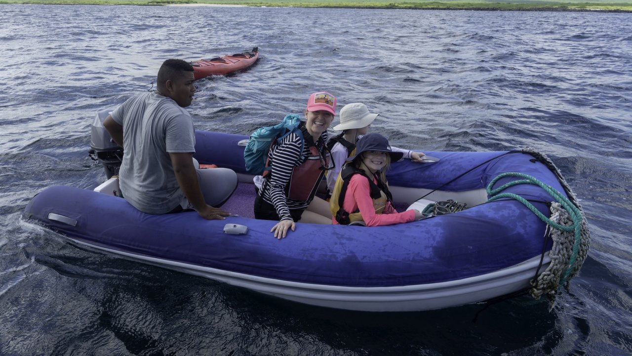 A family with a local guide riding an inflatable boat across blue waters on a Galapagos trip, with kayaks and lush islands in the background.