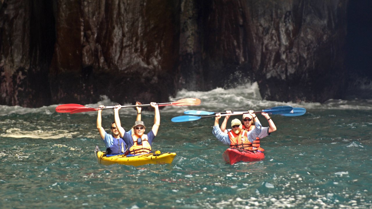 Two tandem kayaks with the paddlers holding their paddles up above their heads while smiling