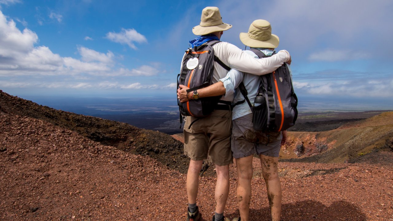 A couple wearing matching backpacks and sun hats overlooking a volcano they hikes up to in the Galapagos