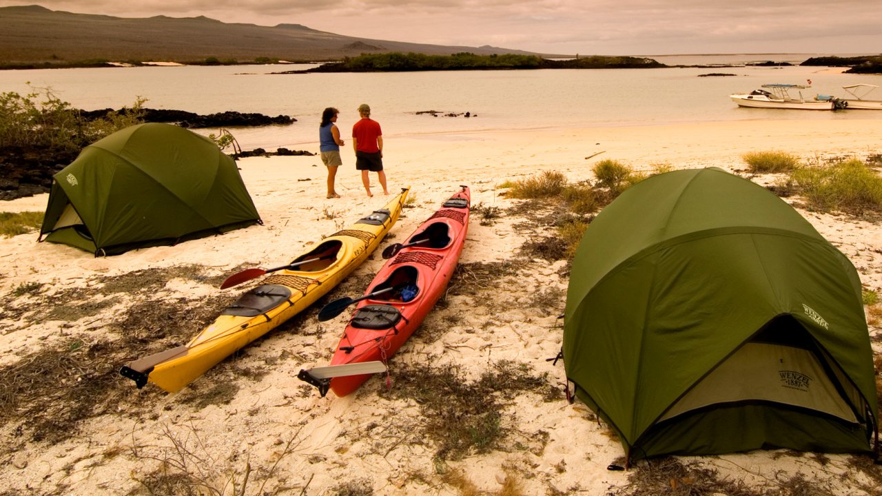 Three sea kayaks in between two green tents on a sandy beach as people walk by with the water in the background