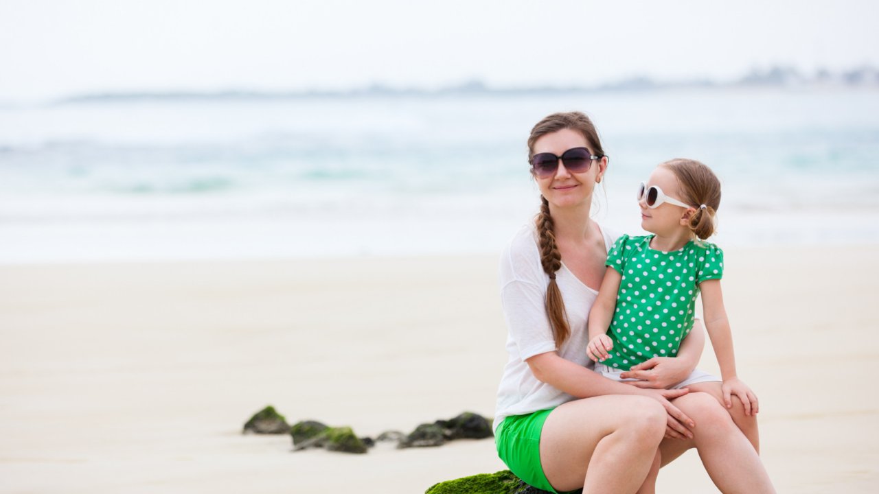 Mom holding her daughter on her lap both wearing sunglasses on the beach with the water behind them in the Galapagos Islands